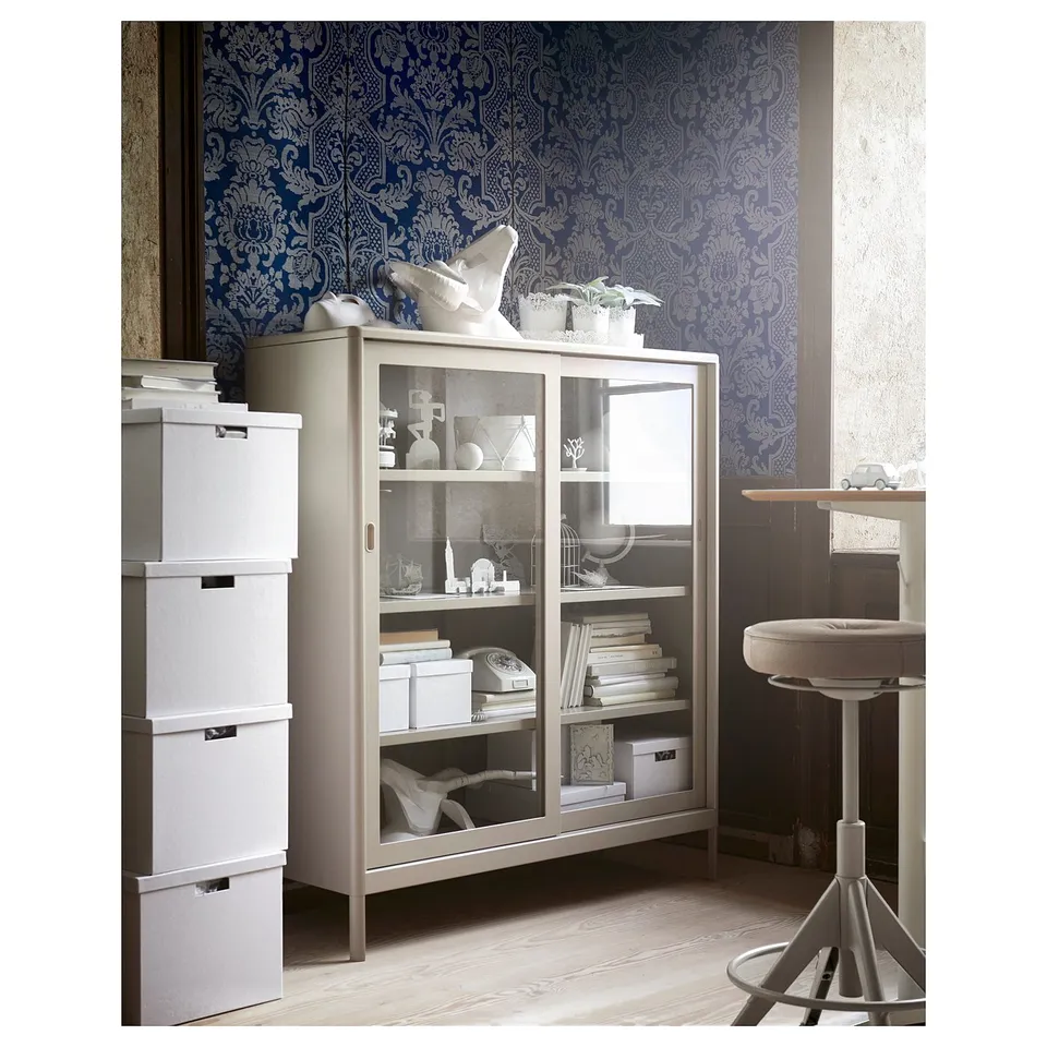 Modern living space with light grey metal cabinet, glass doors displaying decorative items. Blue patterned wallpaper, light wood floor.