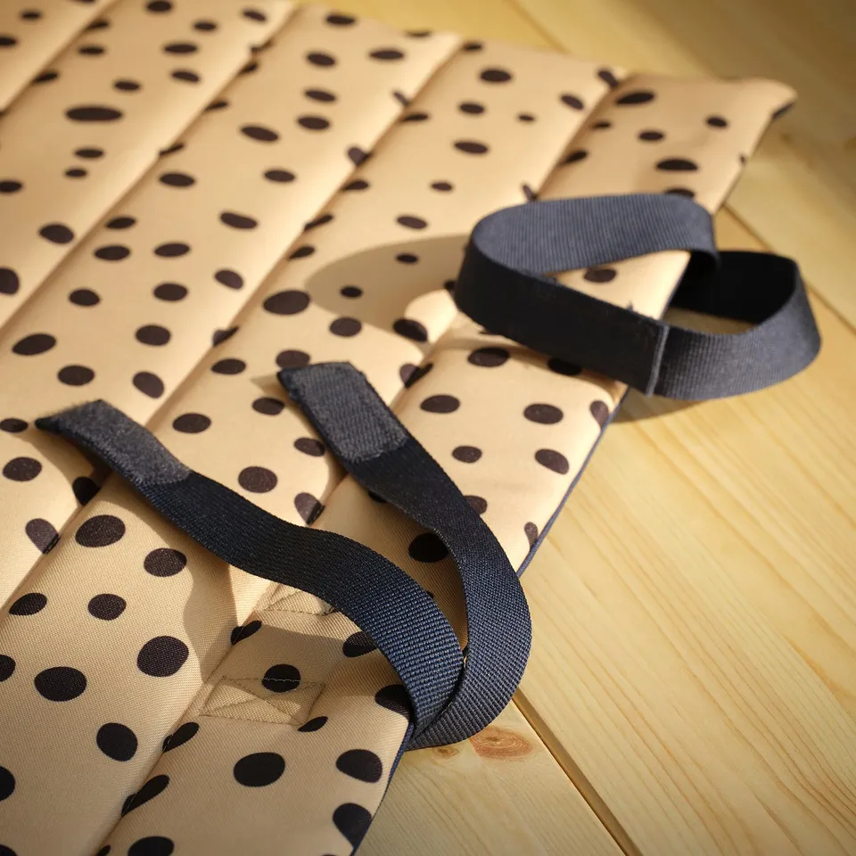 A folded polka dot mat lies on a wooden floor. Two black straps with buckles are attached for carrying. The fabric appears soft and securely stitched.