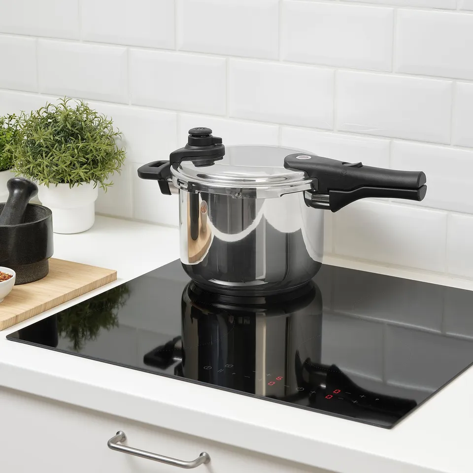 Stainless steel pressure cooker sits on black induction hob, with two black handles and a clear lid.