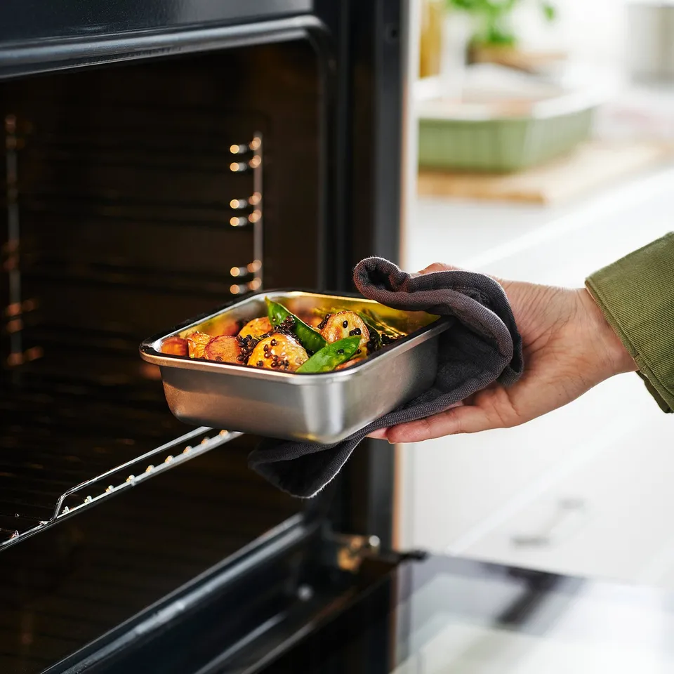Hand holds metal tray with roasted veggies into open oven, demonstrating its oven-safe feature.