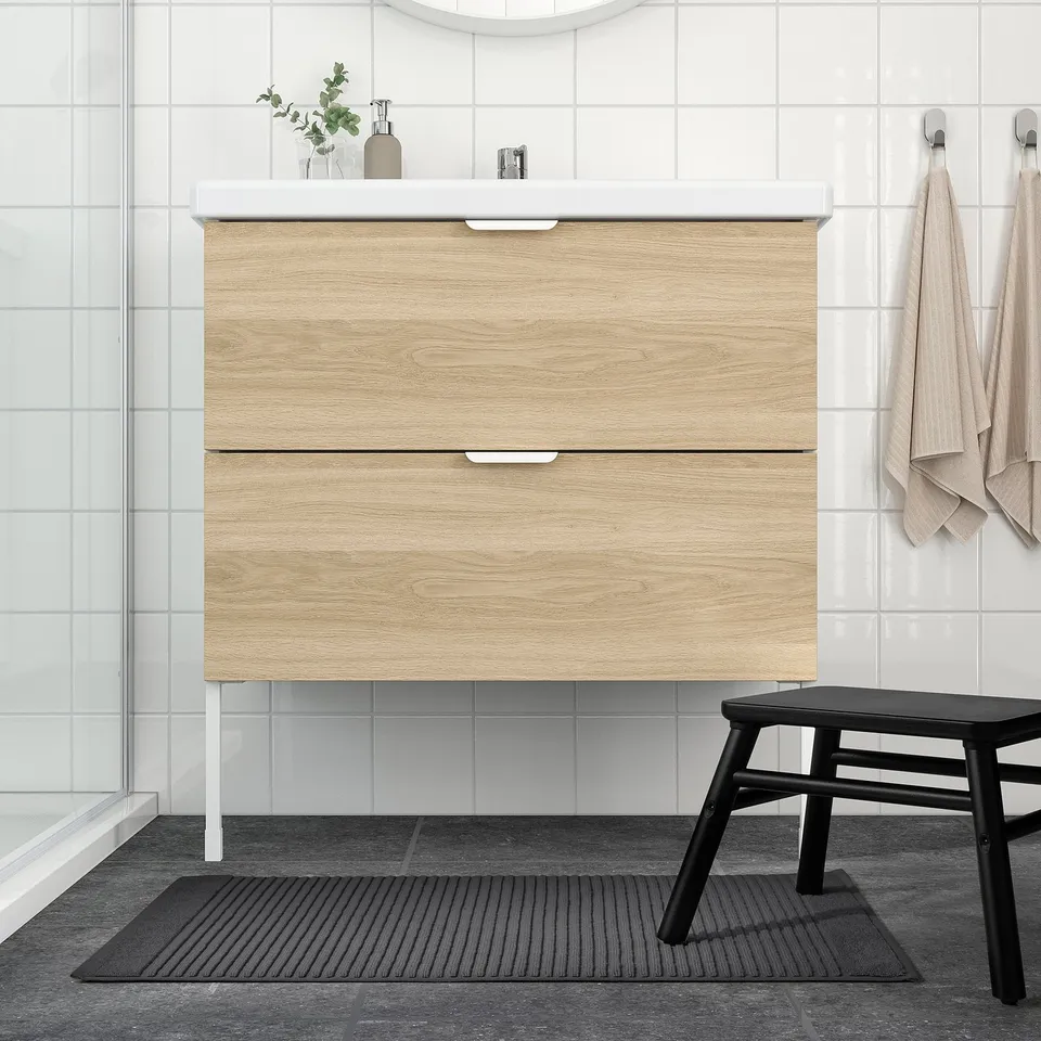 Modern bathroom with light wood vanity, black stool, grey mat, white walls & glass door.