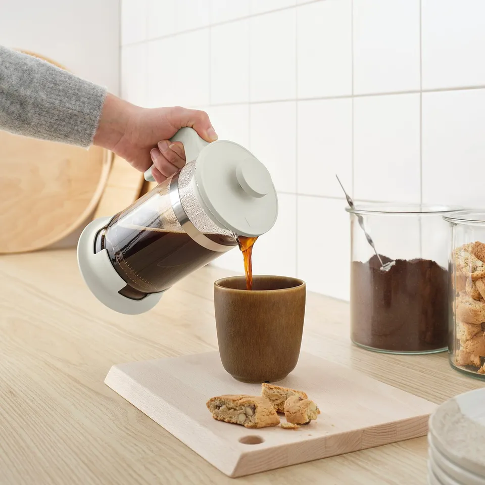 Person pours coffee from a cafetière into a mug on a wooden board with cookies, near jars of cookies and ingredients.