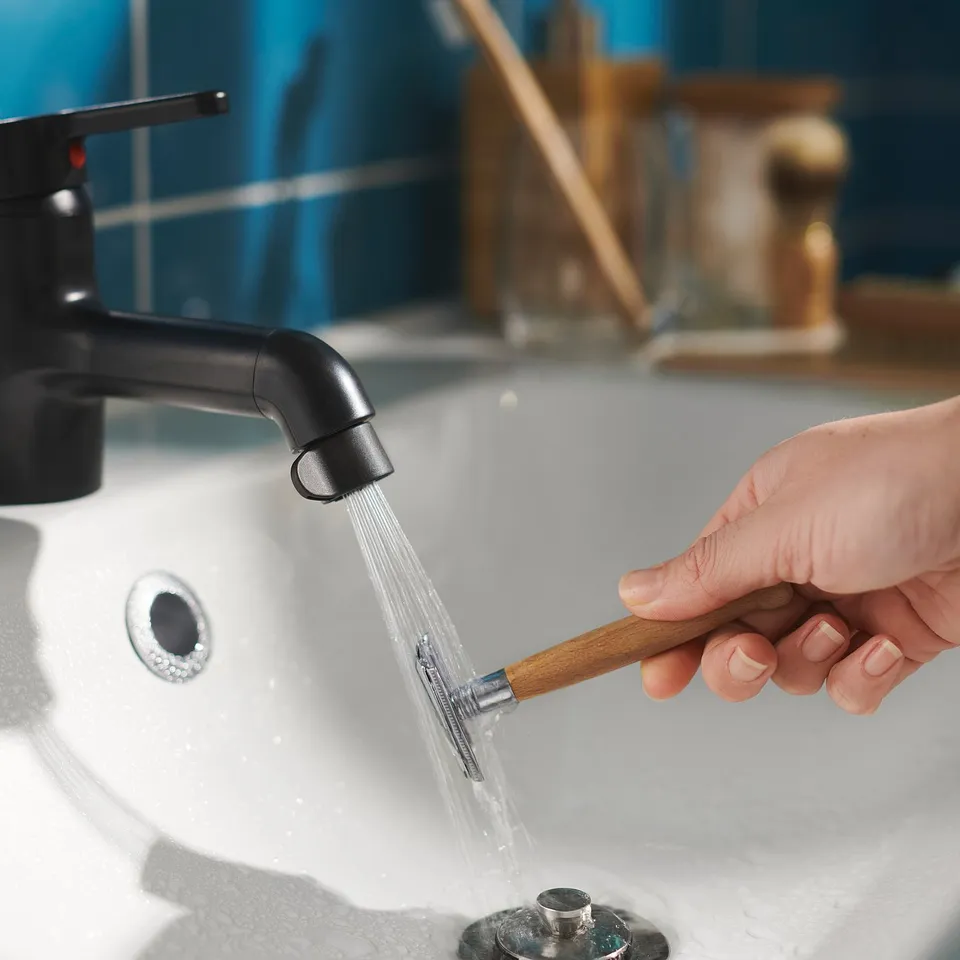 A hand holds a black ÅBÄCKEN water-saving bathroom nozzle over a sink.