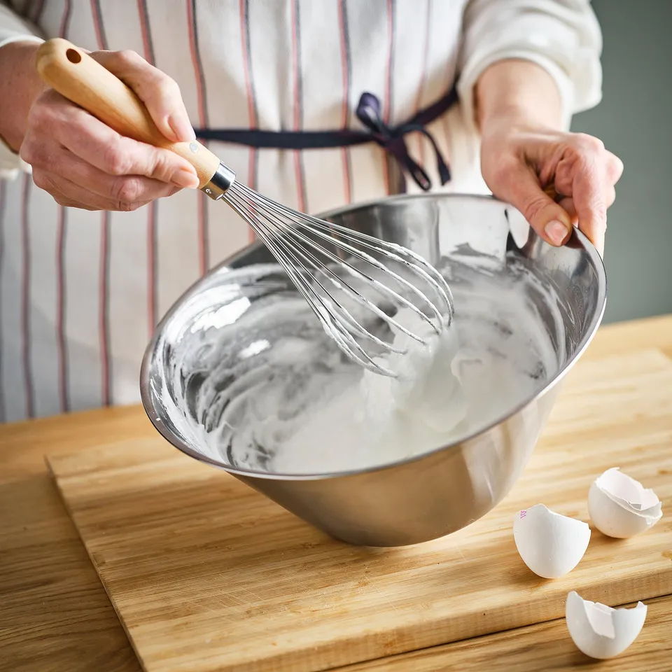 Person whisking mixture in metal bowl with VARDAGEN whisk on wooden table.