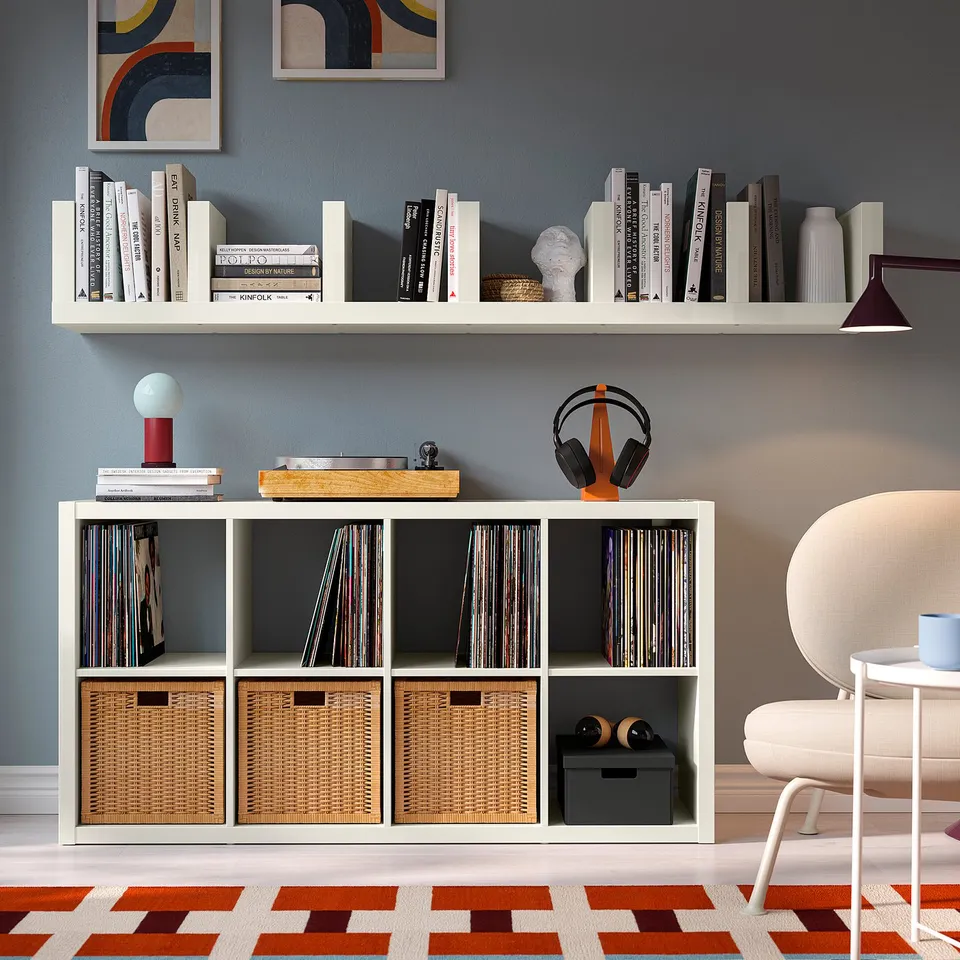 Modern room with LACK shelves holding books and decor. Below, a LACK unit with baskets and records, next to a cosy chair.