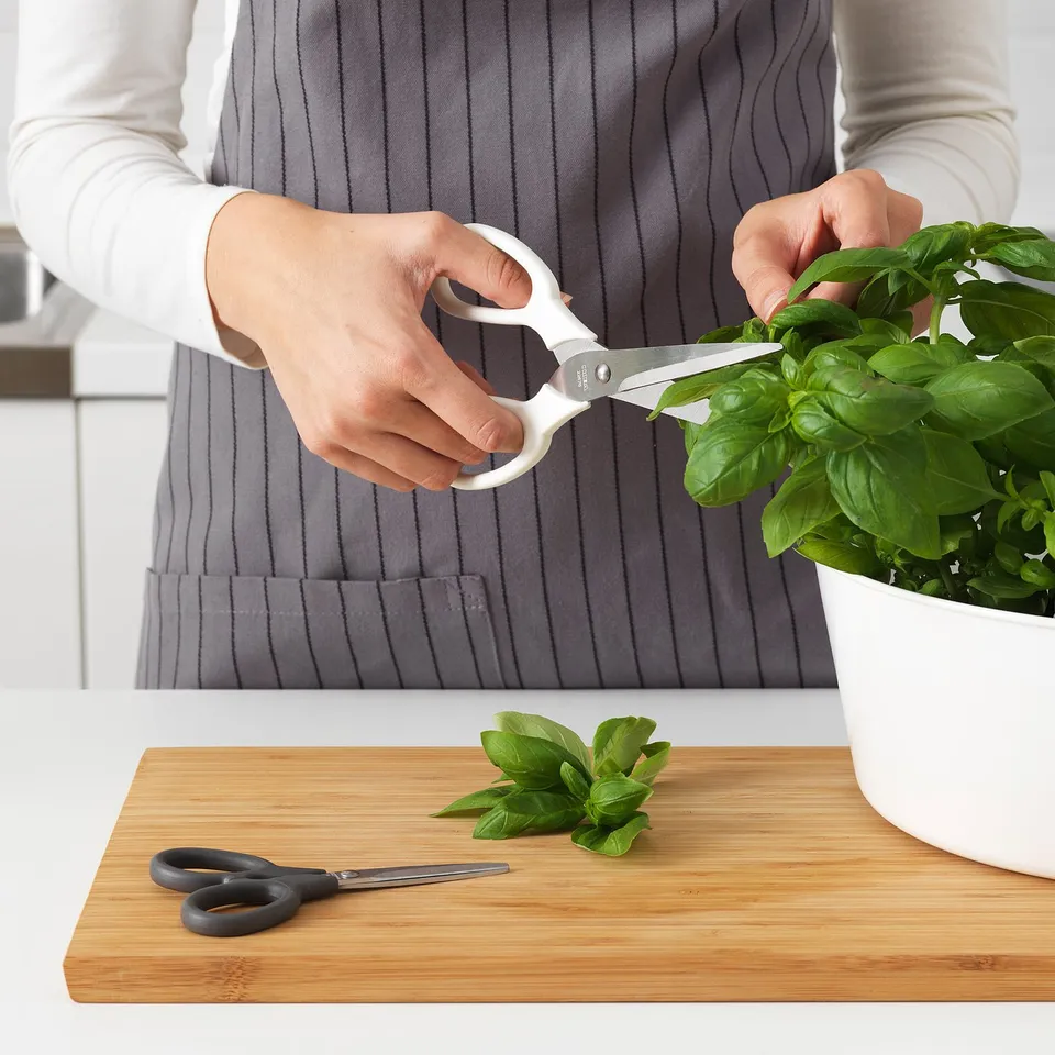 Person trims basil with white kitchen scissors. On the table, theres another pair of scissors and cut basil leaves on a cutting board.