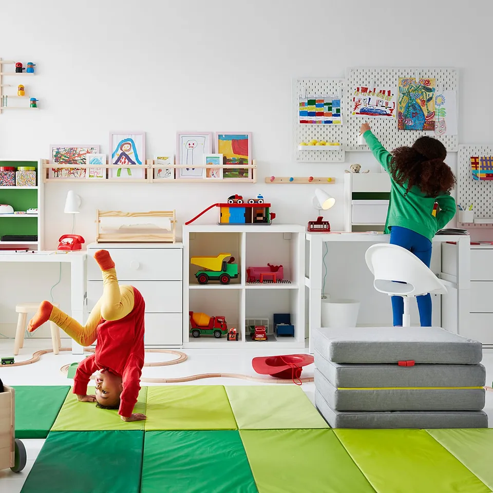 Two children playing in colourful room with LOBERGET chair. One child sits on adjustable white swivel chair, another does handstand on green mat.