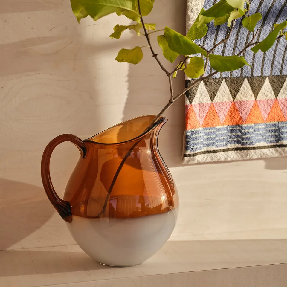 Ommjange amber glass jug with handle, round base, sits on wooden floor, beside plant.