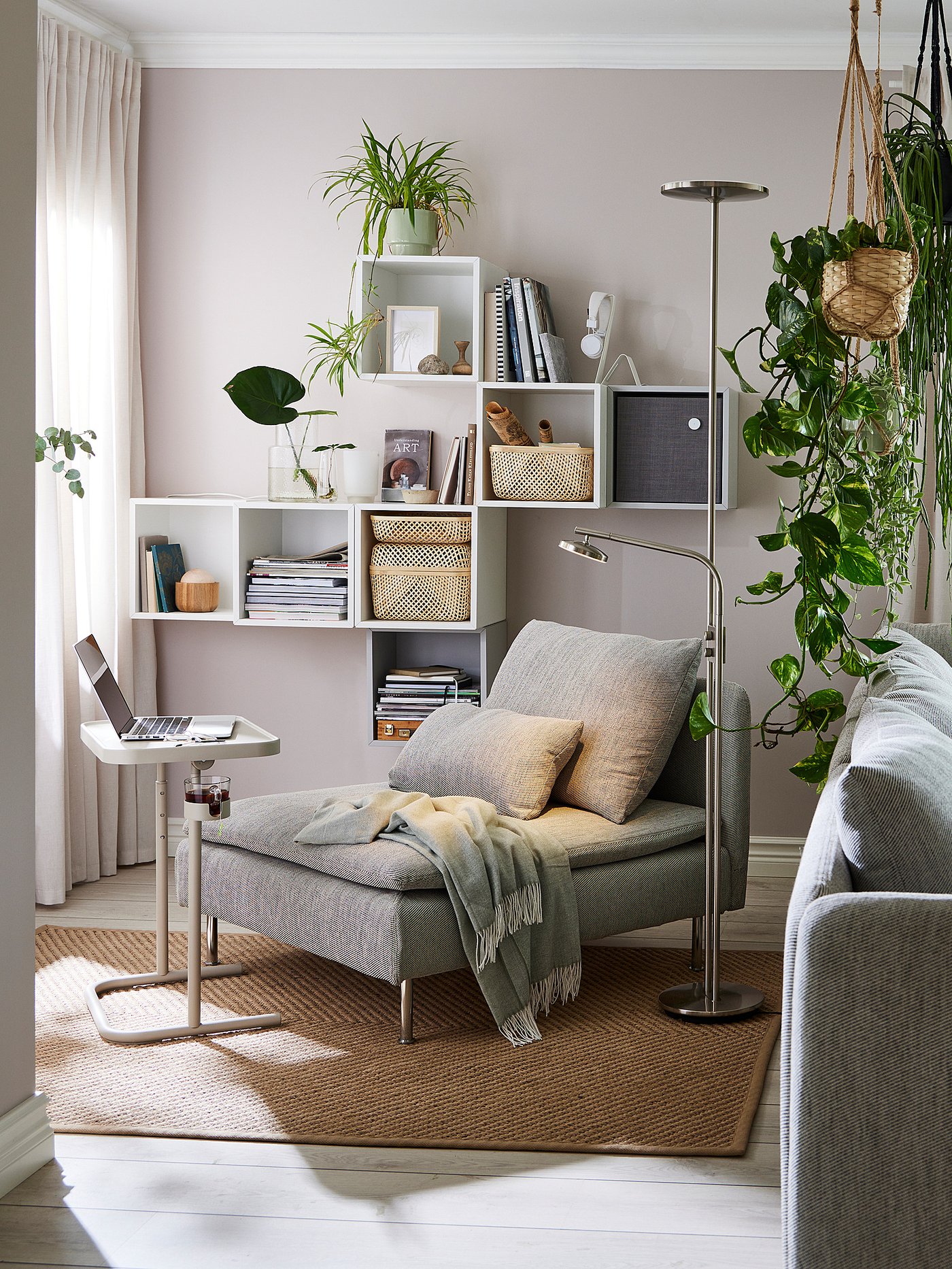 Cosy living room with EKET shelves holding books and plants. Grey chaise with pillows, green blanket, laptop table, and floor lamp.