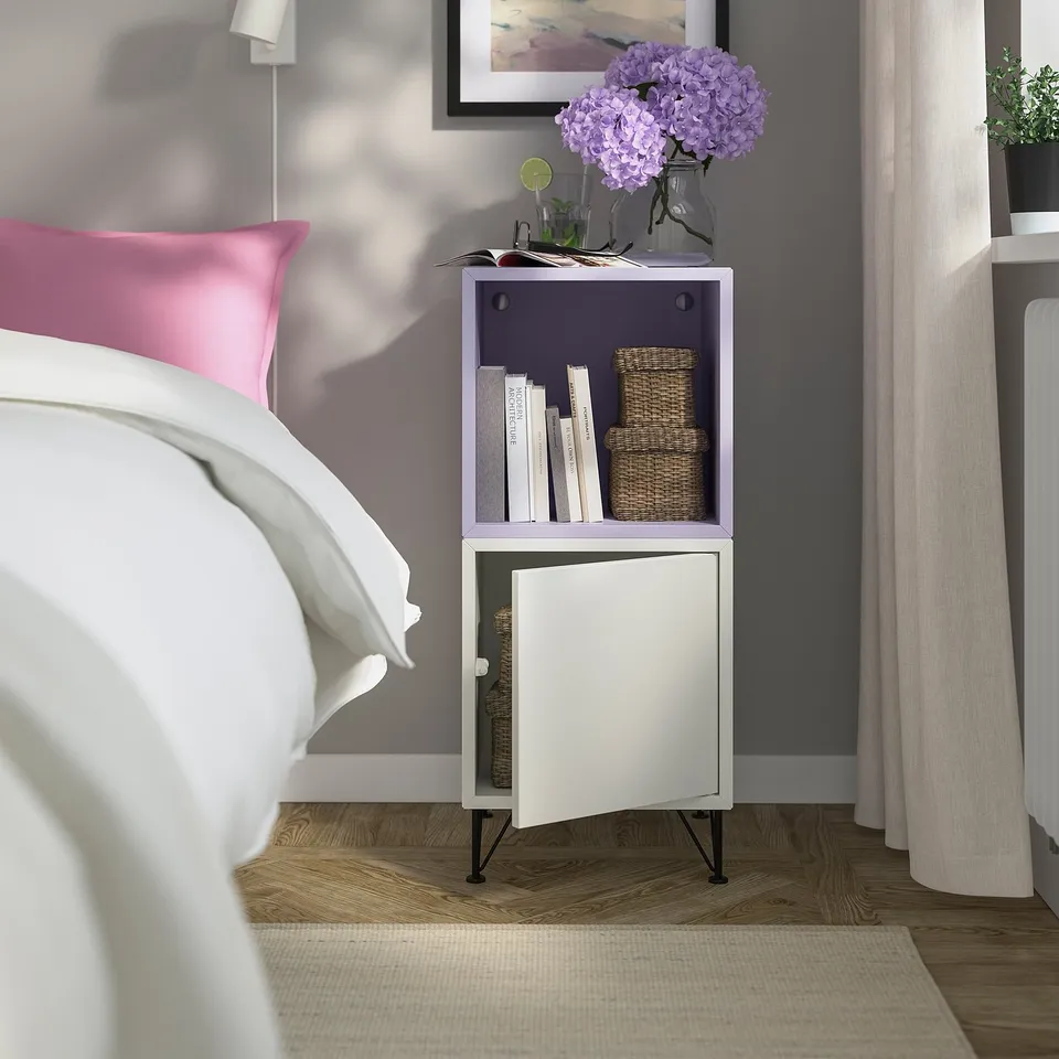 EKET storage unit in white with purple accents sits beside a bed. It holds books, baskets, and a vase with purple flowers.