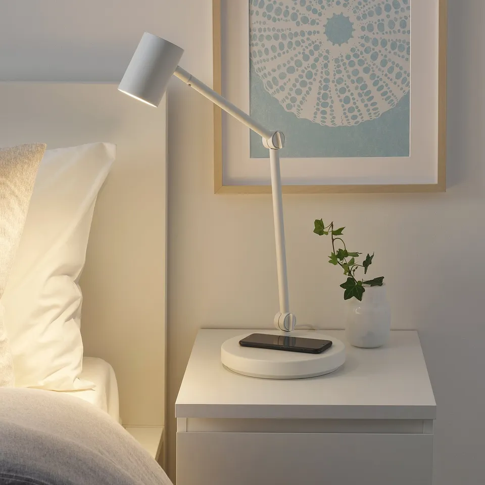 Modern bedside scene with NYMÅNE white lamp, phone, plant, art; lamps adjustable, wireless charging.