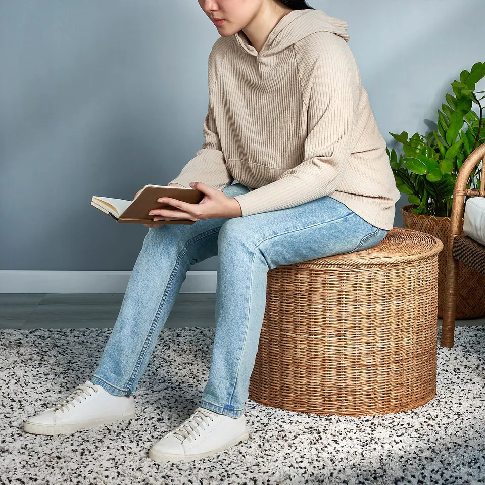 Person sits on wicker stool, reading a book. The stool doubles as storage and extra seating.