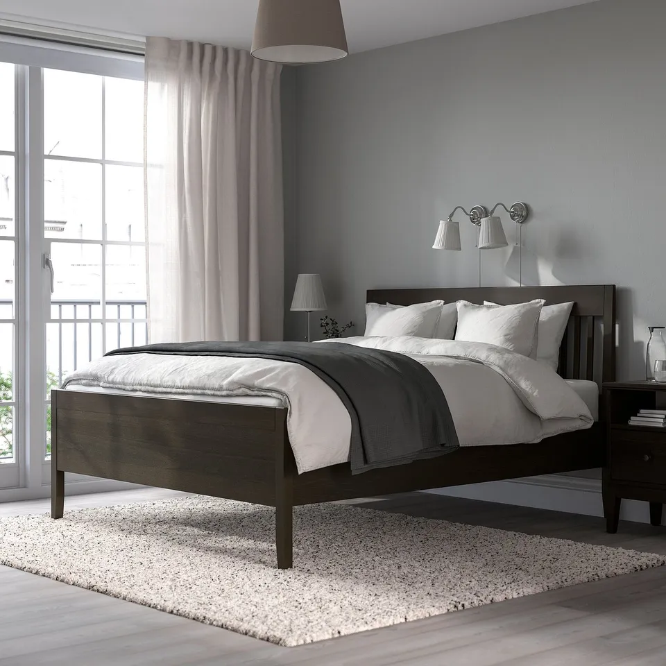 Modern bedroom with IDANÄS dark brown bed, neatly made with grey linens. Large window with curtains and bedside lamps. Rug under bed, cosy setup.