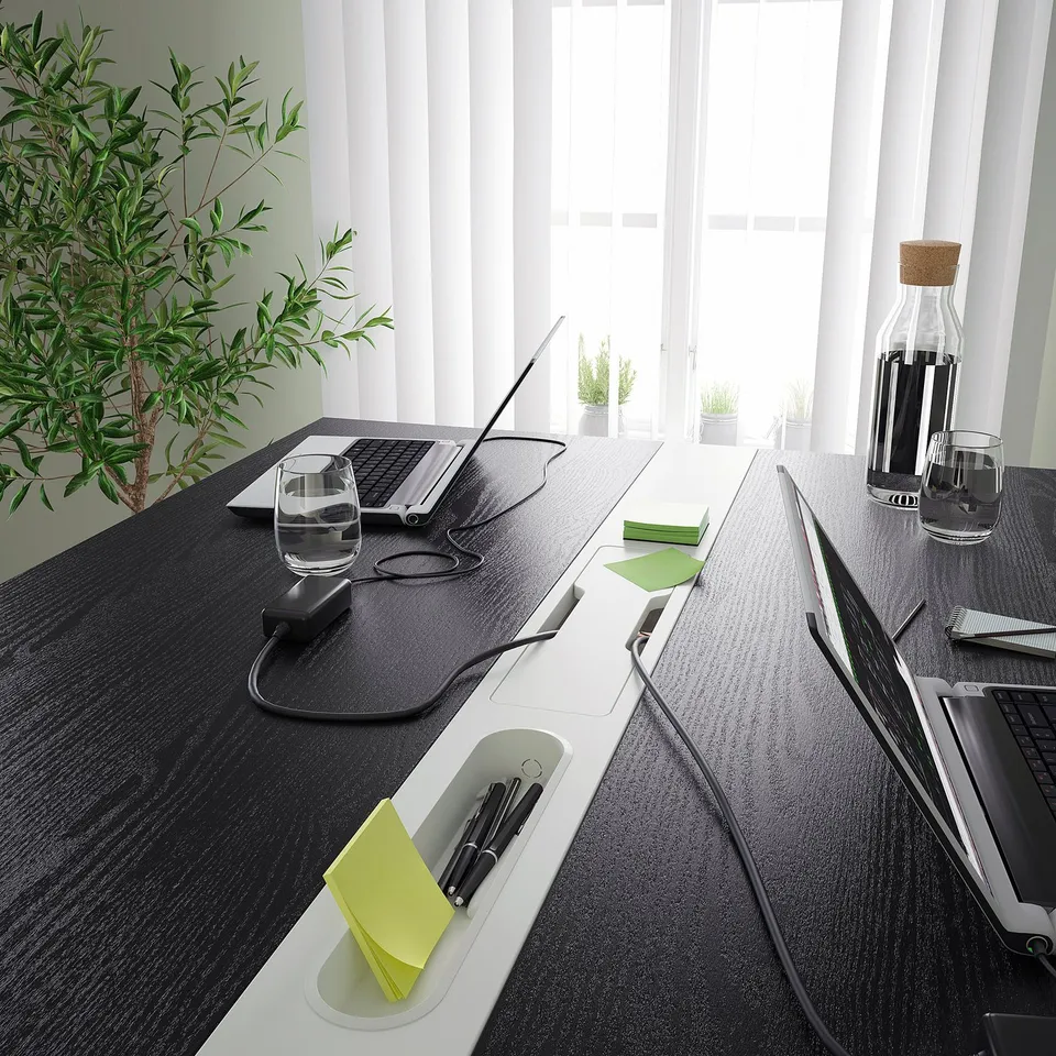 High conference table with cable management and footrest, suitable for sitting or standing, with plants and tech setup.