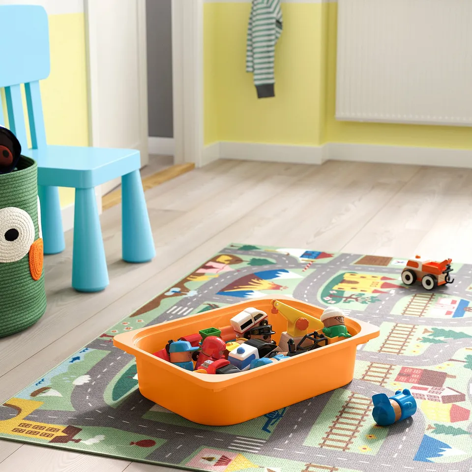 Playful childrens storage bin on colourful rug, holding toy vehicles and figures. Blue chair is nearby.