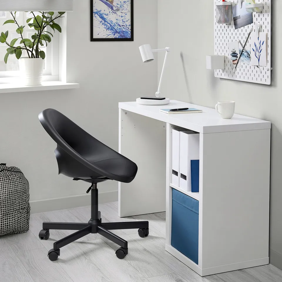 Small white KALLAX desk with blue accents against a wall, featuring a black ergonomic office chair, modern lamp, and decorative plant.
