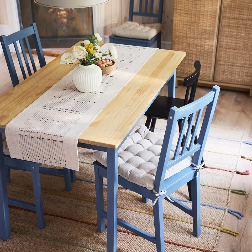 Wooden PINNTORP table with blue chairs, beige table runner, and vase of yellow flowers. Multicoloured rug, ideal for smaller spaces.