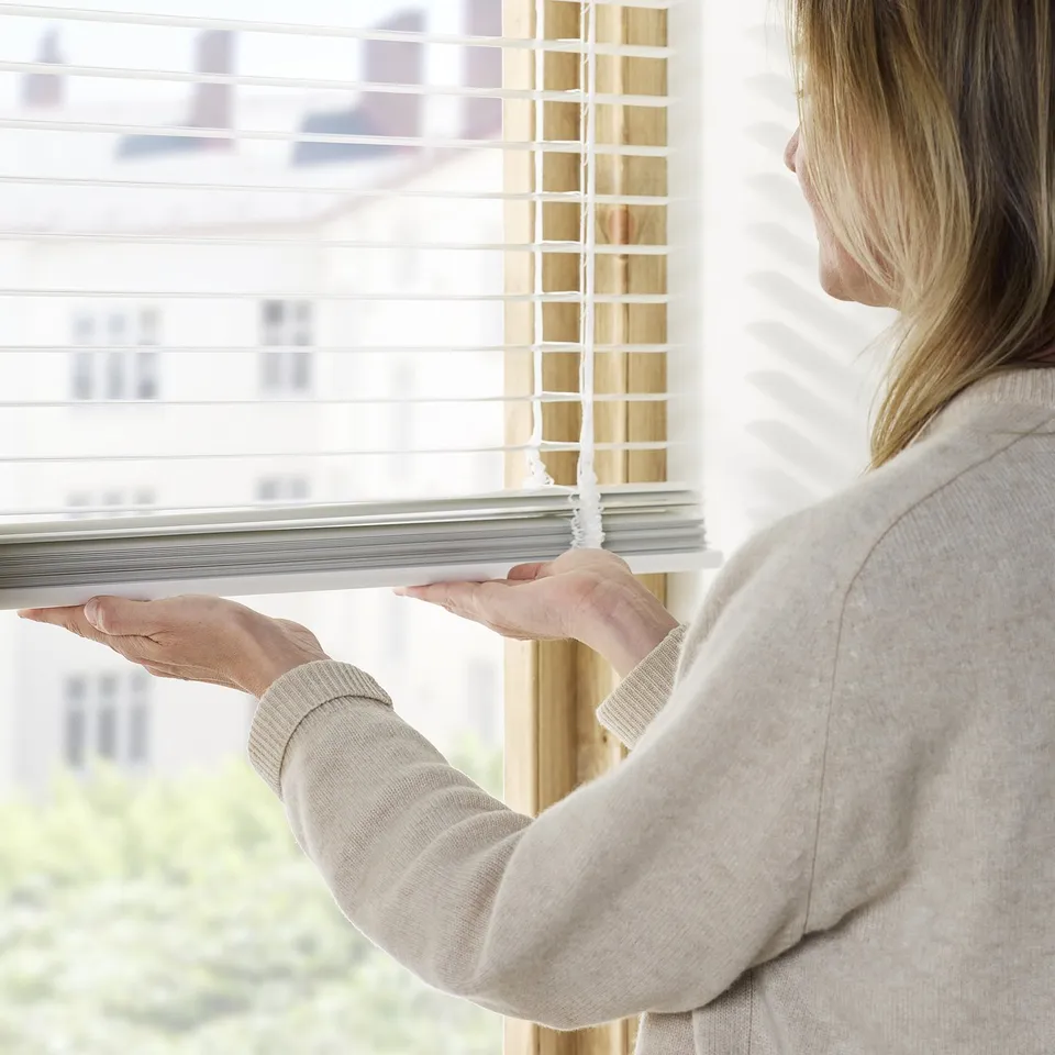 A person adjusts venetian blinds by pulling a cord, controlling light and privacy in a room.
