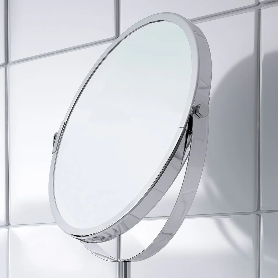 Modern oval magnifying mirror mounted on chrome stand, with sleek design suitable for shaving or makeup. Reflects neatly in clean, white-tiled room.