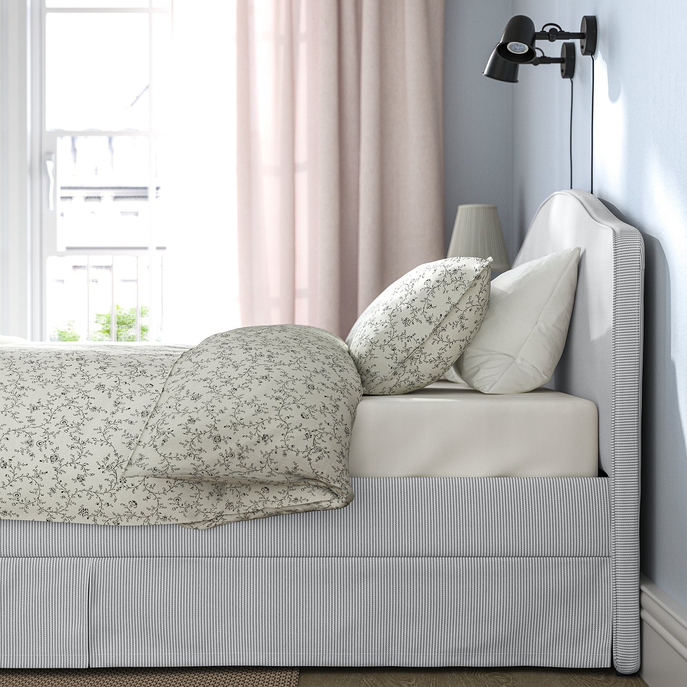 Bed with grey-white striped cover and floral patterned blanket. Includes decorative pillows and wall-mounted reading lamp.