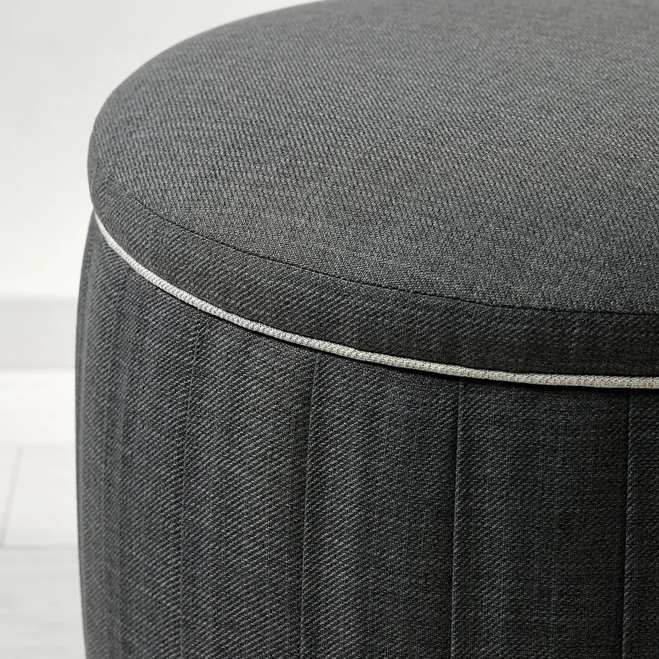 Dark grey upholstered pouffe with neat vertical pleats and piping, showcasing quality craftsmanship.