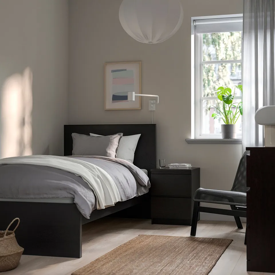 Minimalist bedroom with grey bed, black drawers, abstract art, pendant lamp, plant, and rug.