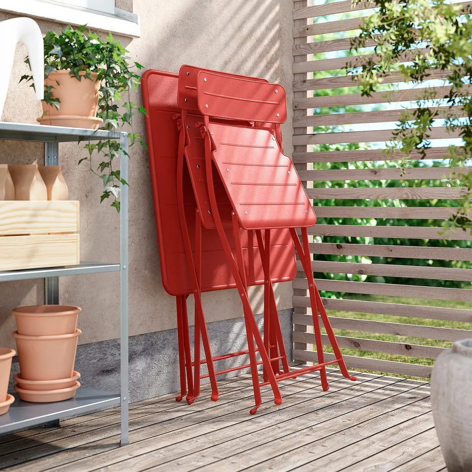 Three bright red SUNDSÖ folding chairs stacked neatly outdoors near a wooden fence and potted plants.