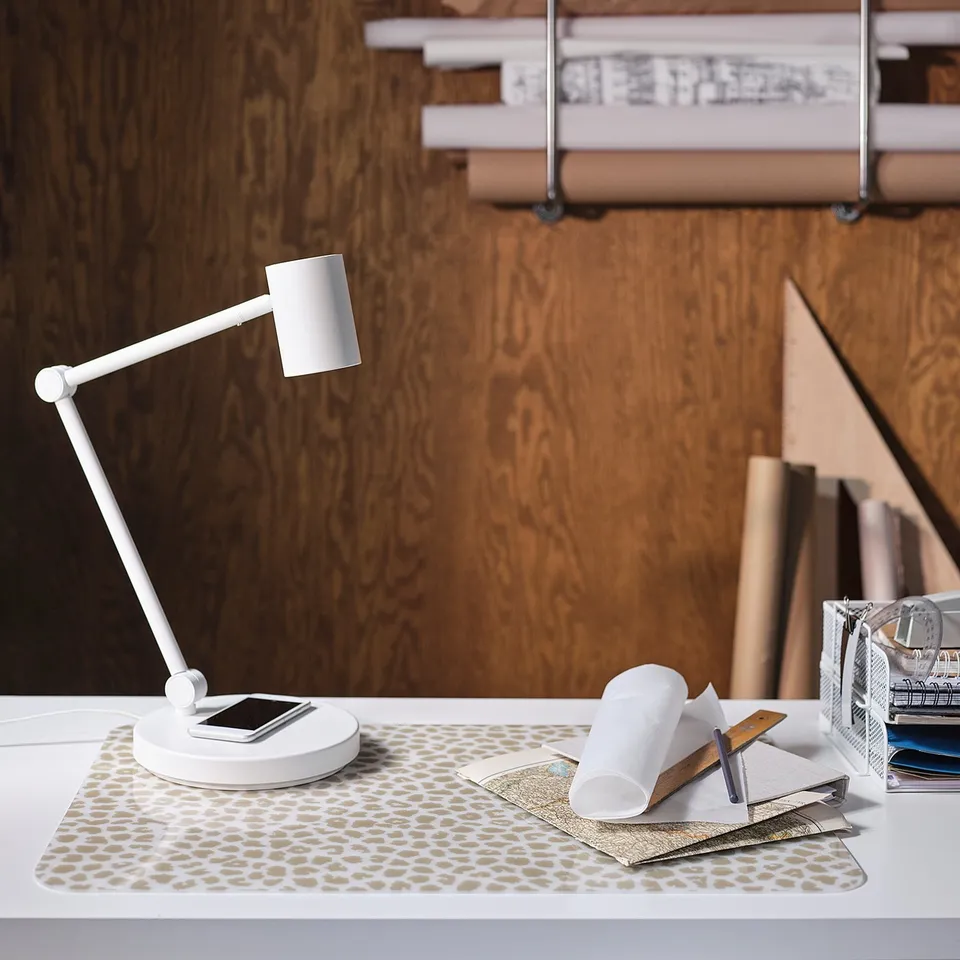 Modern white desk lamp with adjustable arm and wireless charging. Leather patterned mat, books, and accessories on table.