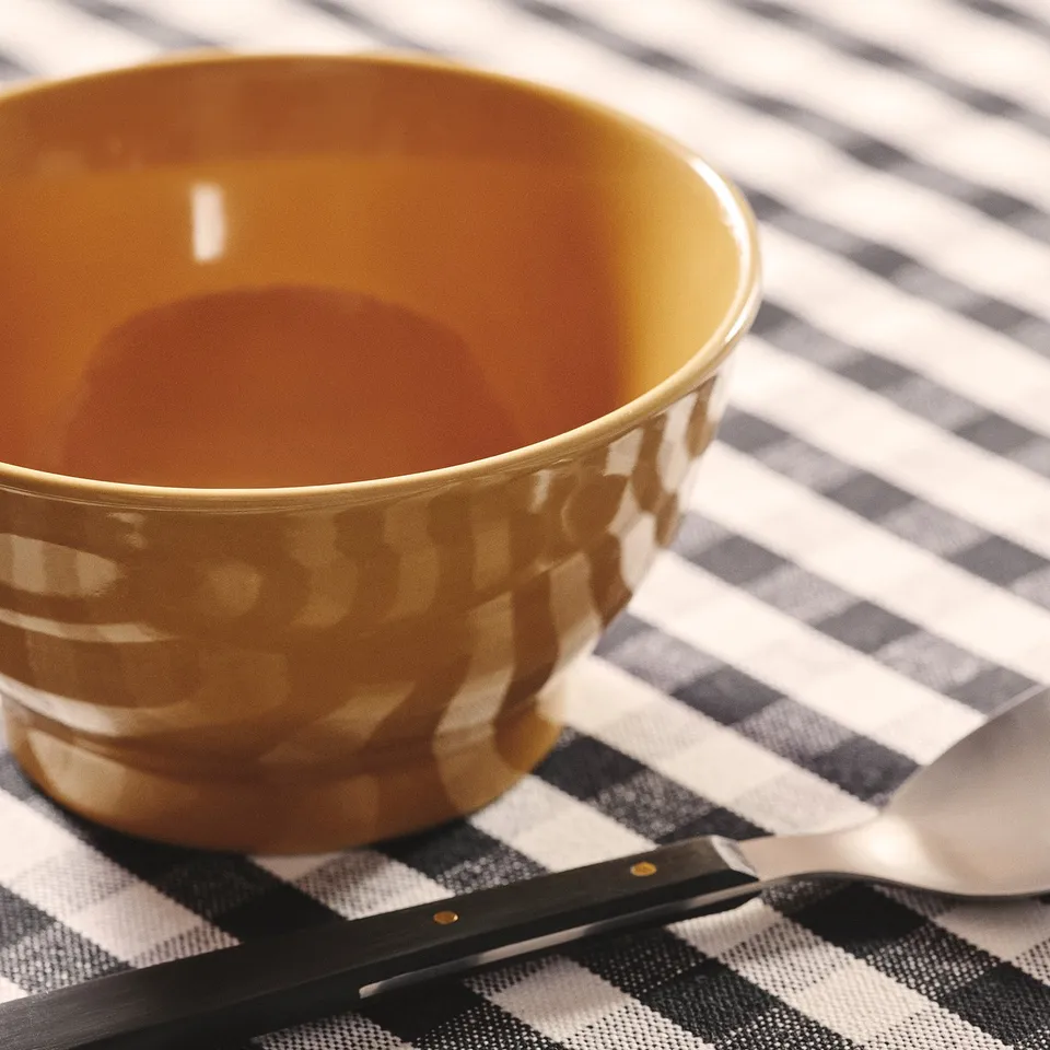 Empty, rustic, ochre-yellow stoneware bowl on chequered tablecloth, with knife and spoon.