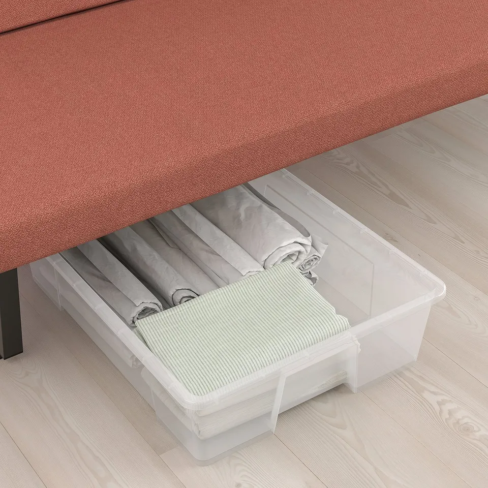 Clear plastic storage bin under red sofa containing folded bed linens.