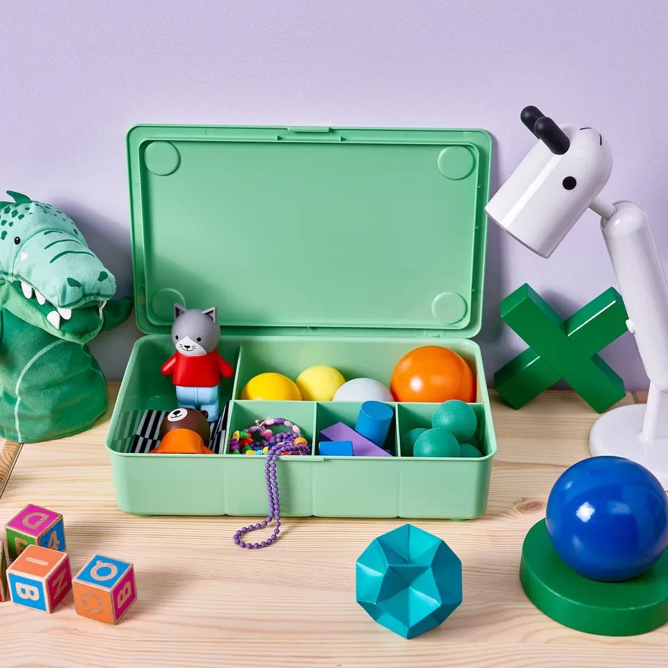 Green toy box with grey cat toy and colourful balls; wooden blocks, dinosaur plush, and robot lamp nearby.