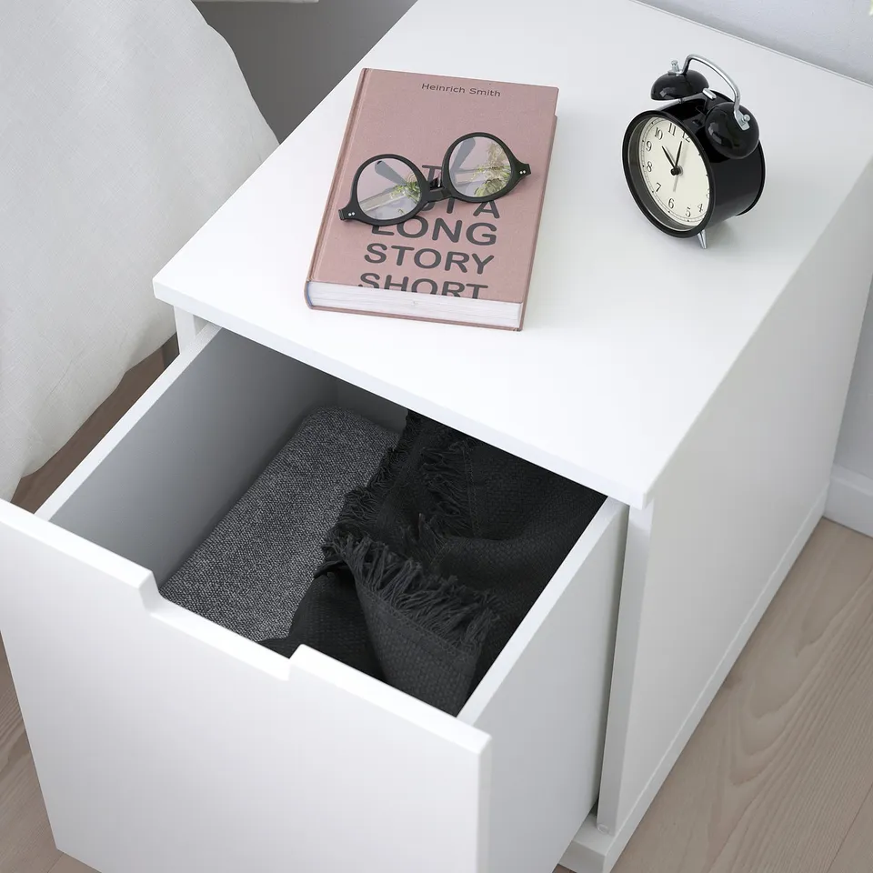 White bedside table with one open drawer revealing folded black blanket. Top has book and alarm clock.