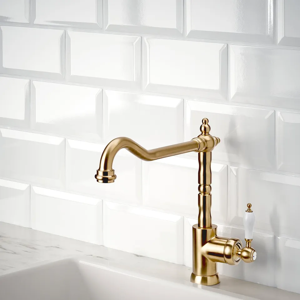 Golden kitchen tap with curved spout and side handle against white tiles.