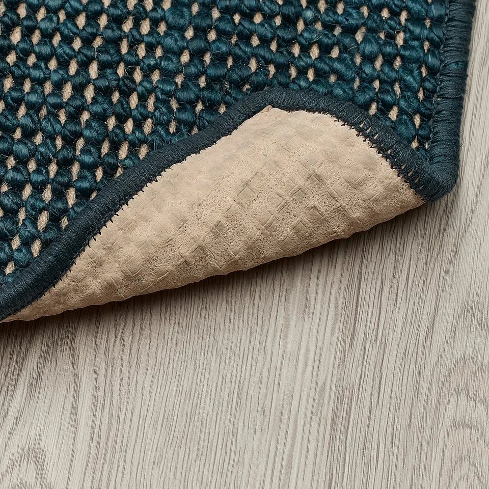 A blue jute door mat with a latex backing lies on a wooden floor. The backing prevents slipping and grips the floor effectively.