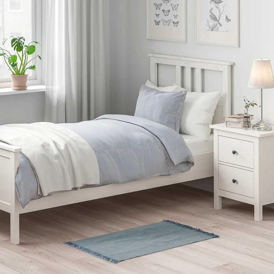 A cosy bedroom with a white bed, blue rug, bedside table with lamp, and butterfly art.