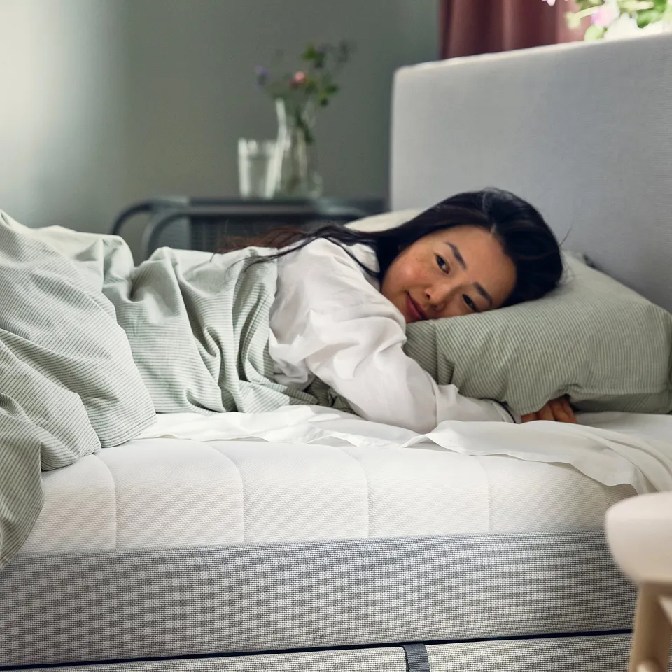 Person sleeping on white foam mattress in cosy bedroom. Laptop and flowers nearby. Comfortable and serene setting.