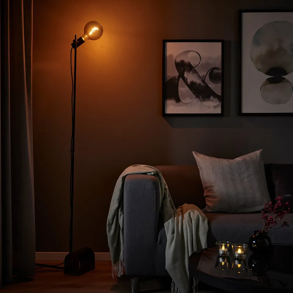 Cosy living room with low-lit HÅRSLINGA floor lamp, blanket, and pillows. Wall art and candles nearby.