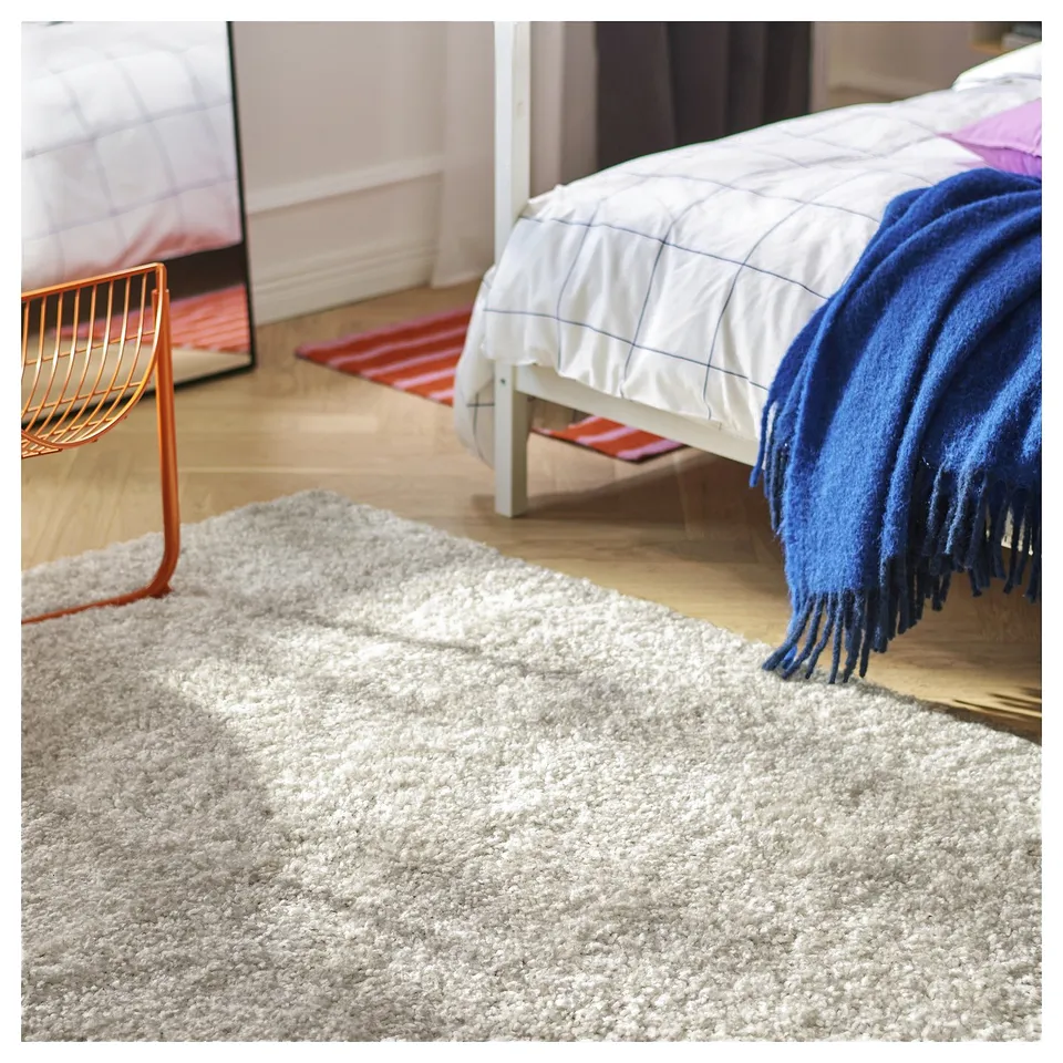 Cosy bedroom with white bed, blue blanket, red rug, and orange chair on fluffy white rug.