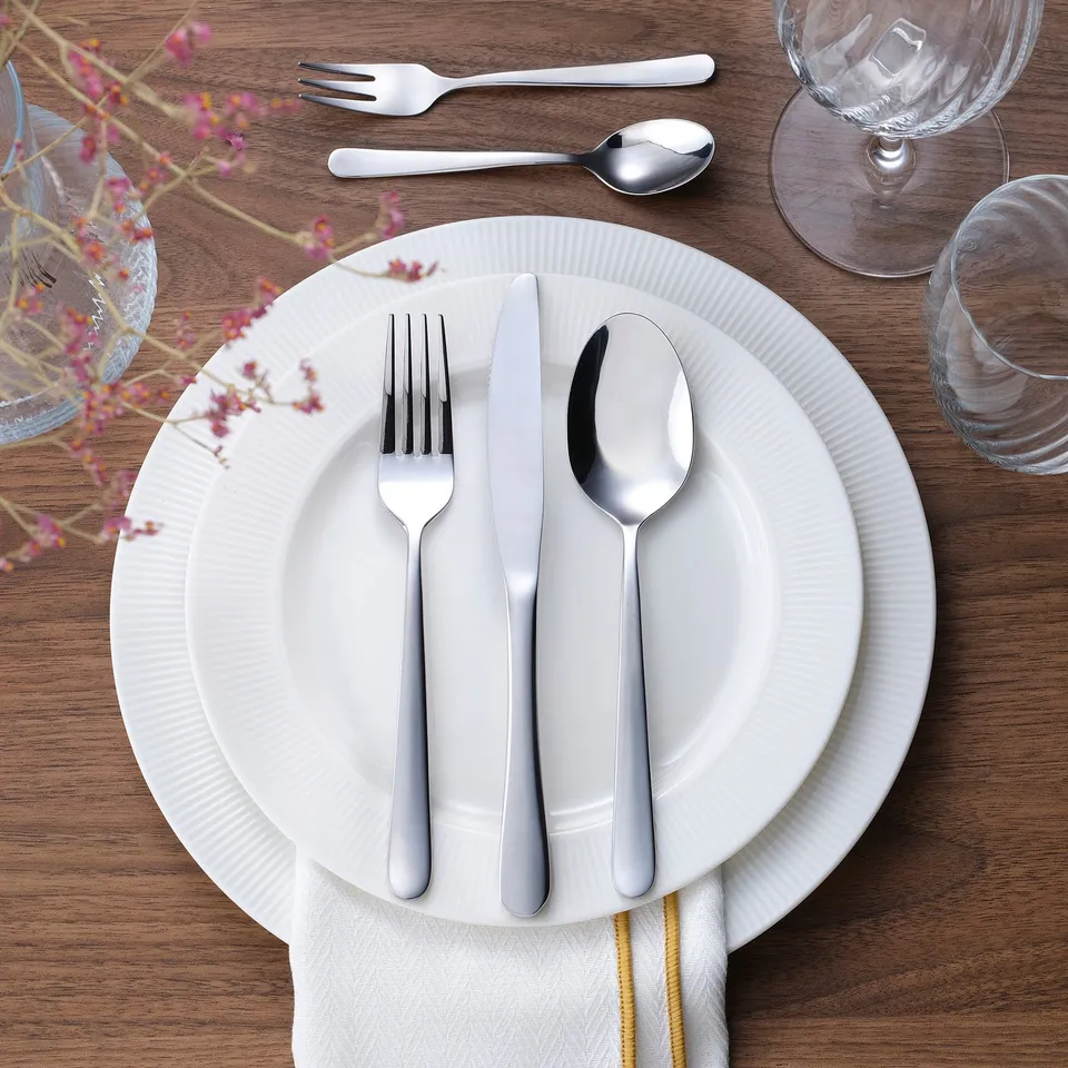 Elegant MARTORP cutlery: fork, knife, spoon on white plate, wooden table, glassware. Classic design, stainless steel, neatly arranged.