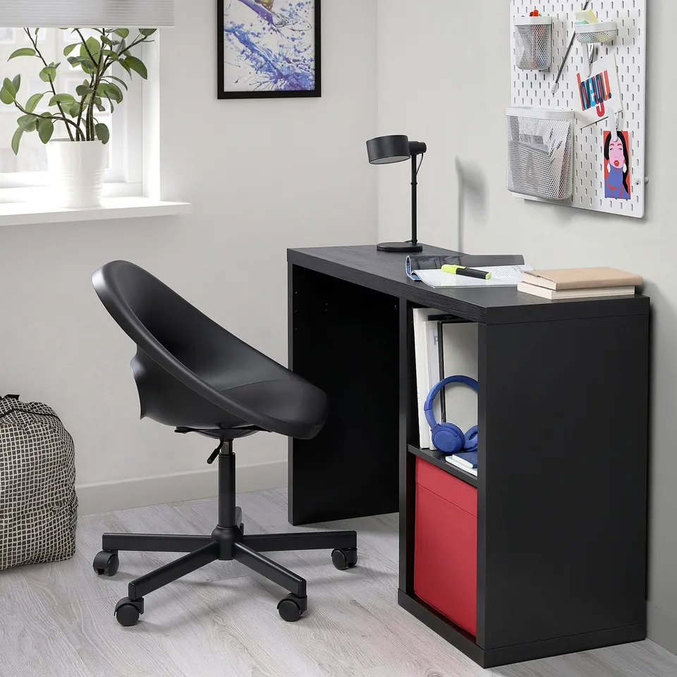 Small black office desk with red storage bin, next to black swivel chair. Brightly lit, with minimal decor.