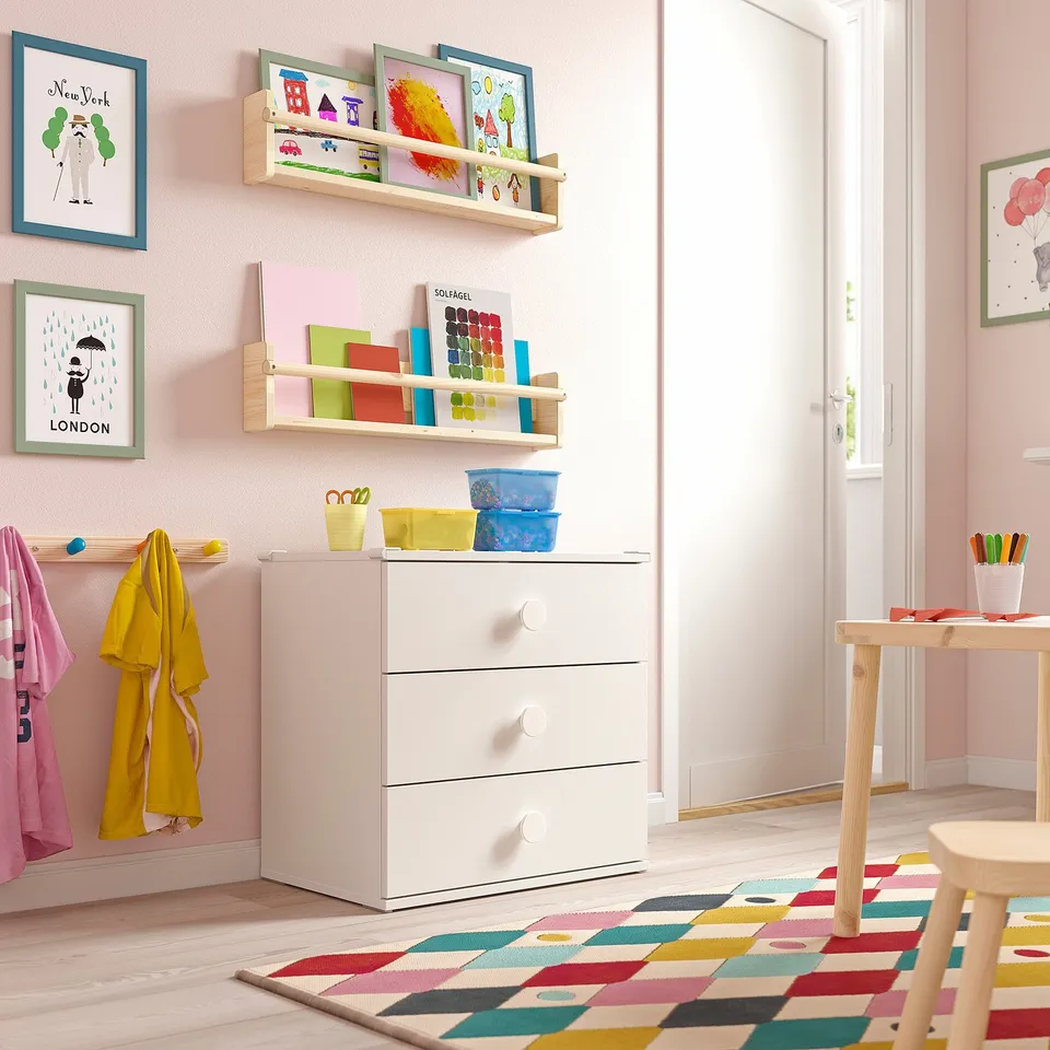 Bright childrens room with GODISHUS white chest, colourful rug, wooden table, and hanging artwork. Drawers are labelled with stickers.