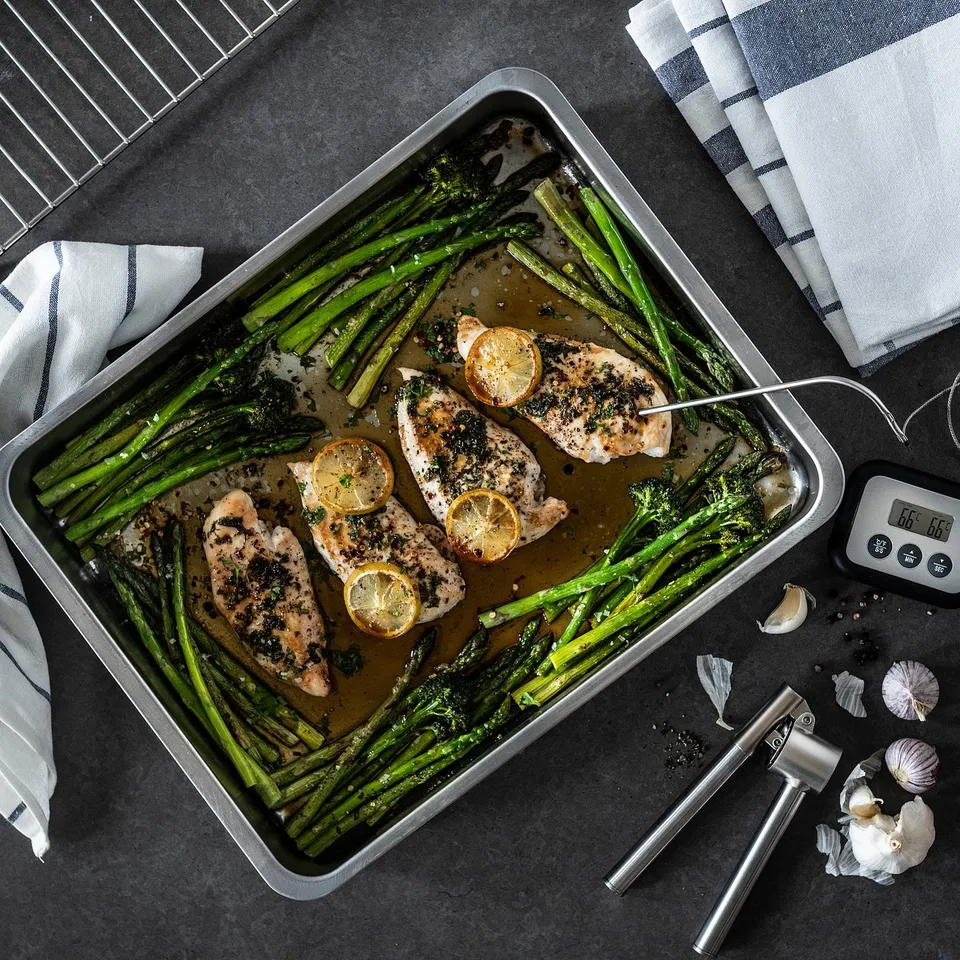 Silver stainless steel roasting tray with chicken, asparagus, lemons, garlic, tongs, and thermometer.
