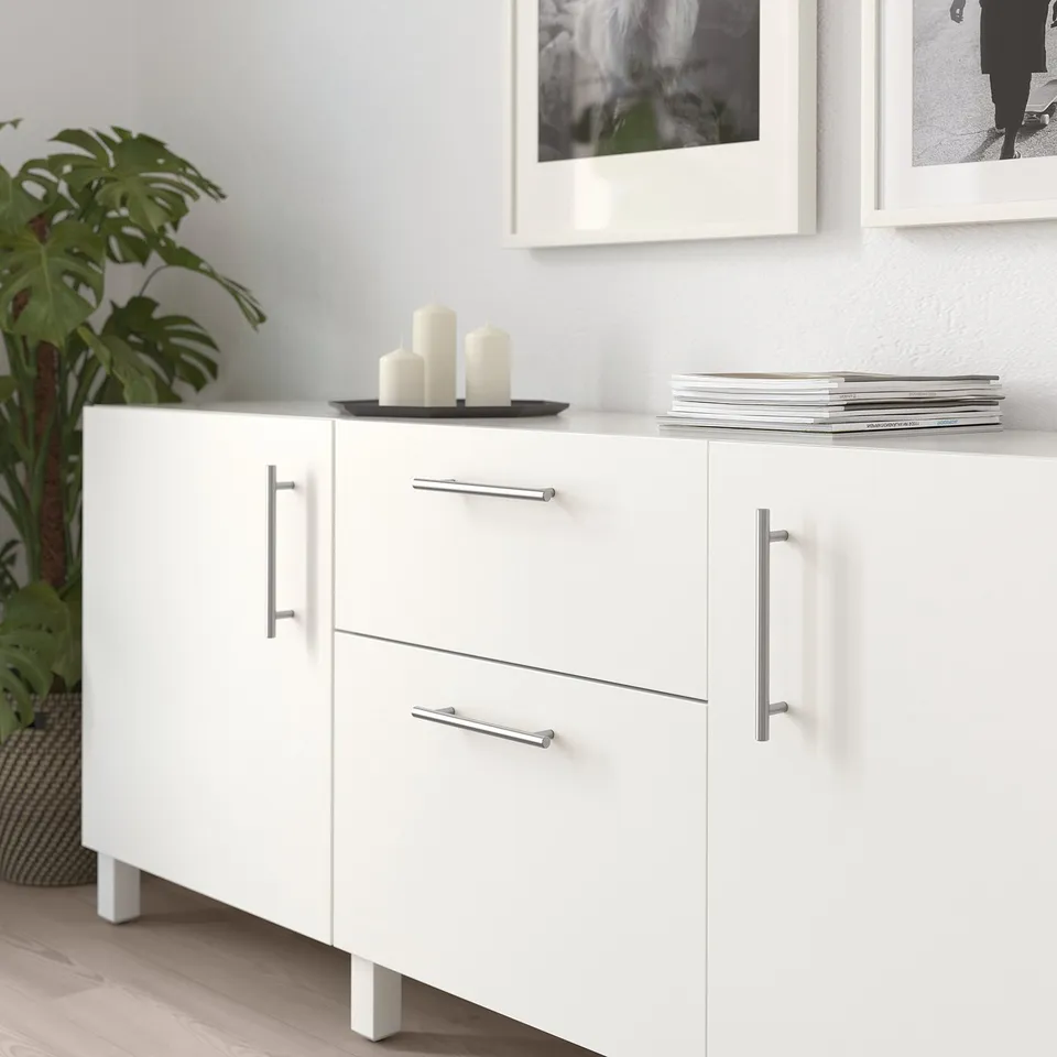 White cabinet with stainless handles, candles, magazines, drawers, and plant. Modern minimalist.