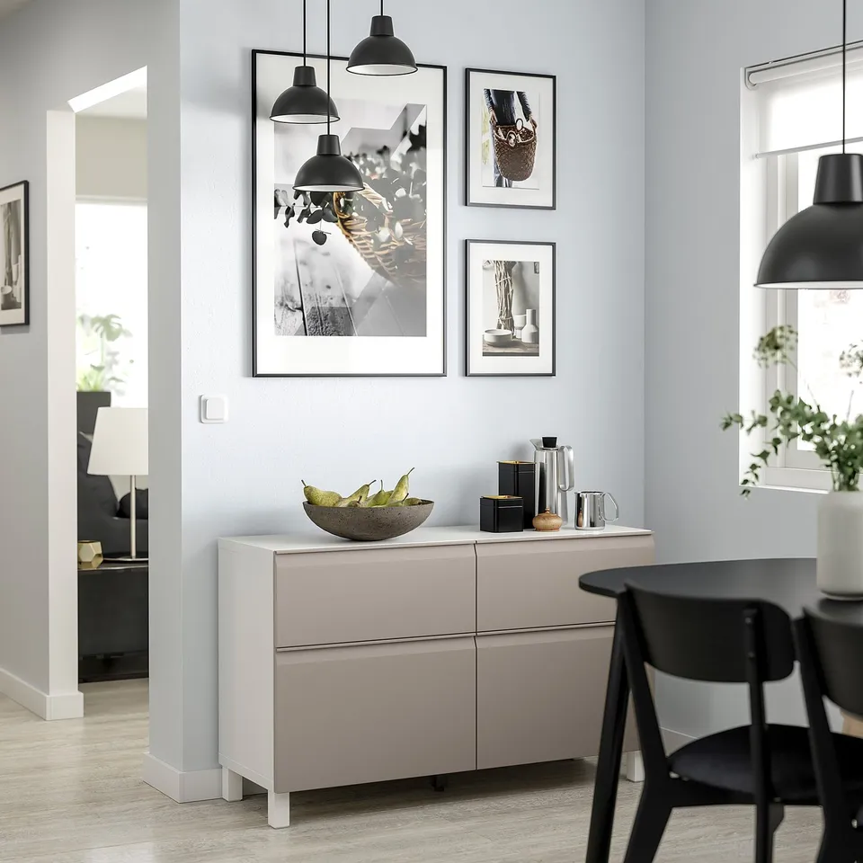 Modern dining area with light grey BESTÅ sideboard, black chairs, and pendant lamps. Artwork and fruit bowl on the sideboard.