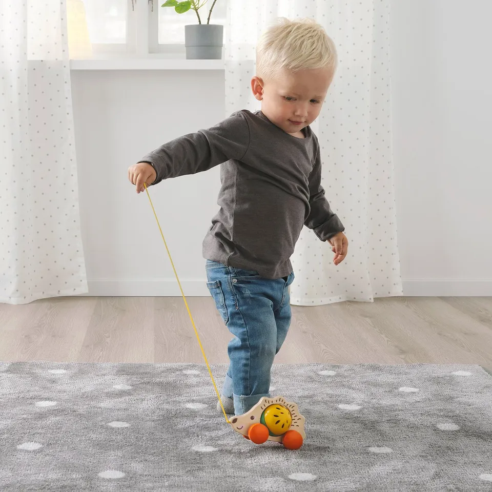 Blonde toddler plays with wooden hedgehog pull toy. Child holds string, pulling toy across speckled rug.