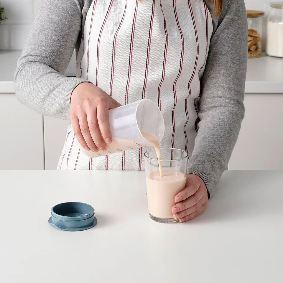 Person pouring liquid from a clear container into a glass on a white counter. A blue lid sits nearby.