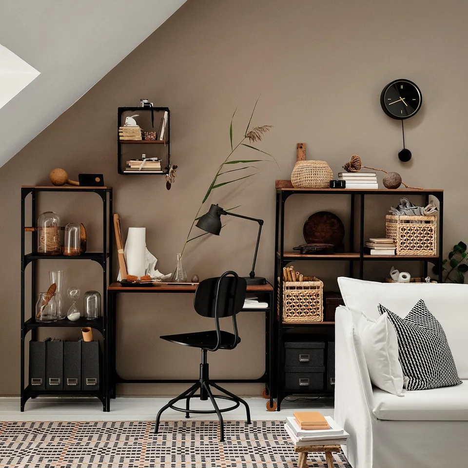 Modern home office FJÄLLBO black metal and light wood shelves, a black office chair, and a white sofa with black and white pillows.