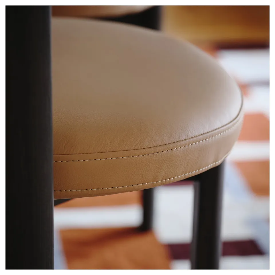 Close-up view of a modern barstools round, beige leather seat. Visible stitching shows high-quality craftsmanship. Black metal legs support the seat.