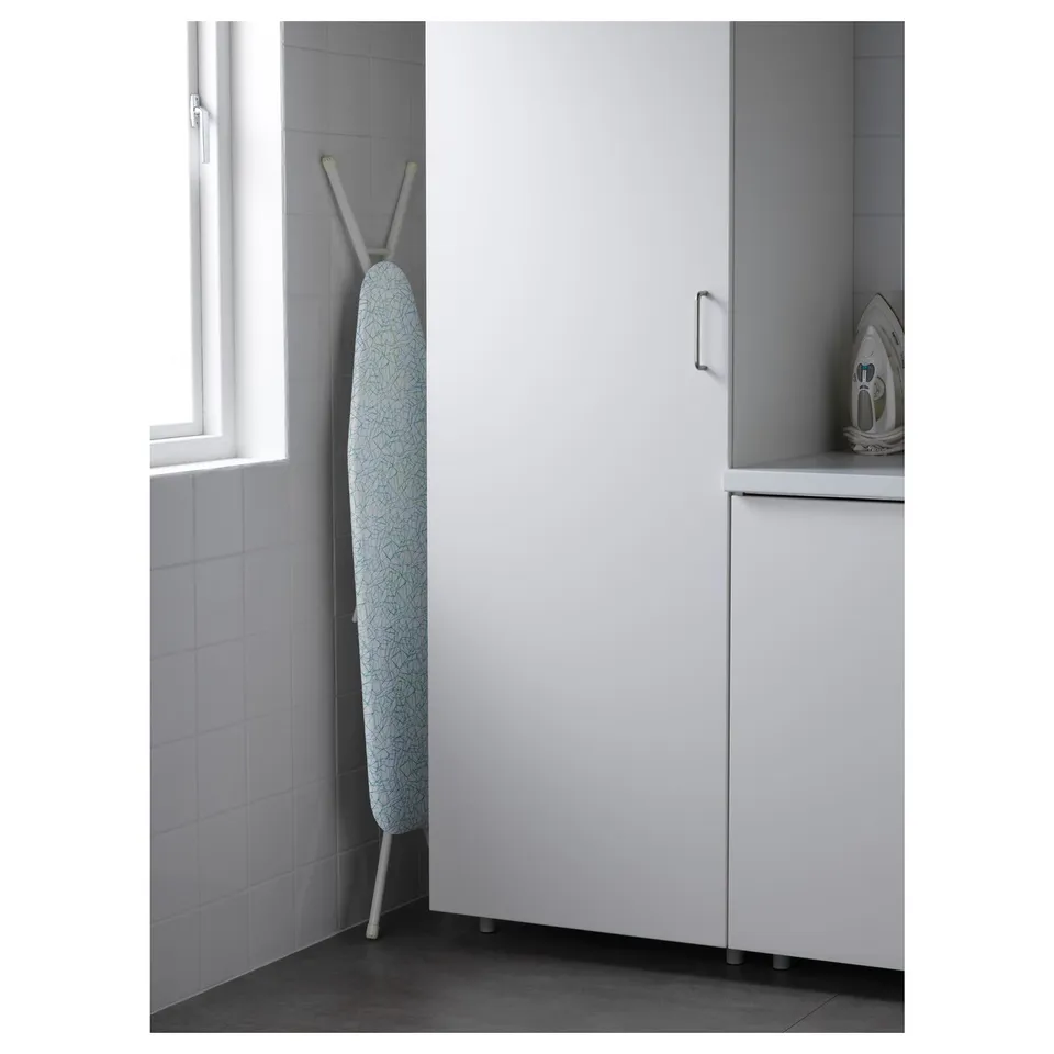 Adjustable ironing board in laundry room corner, next to tiled wall and window, with steel frame for stability.