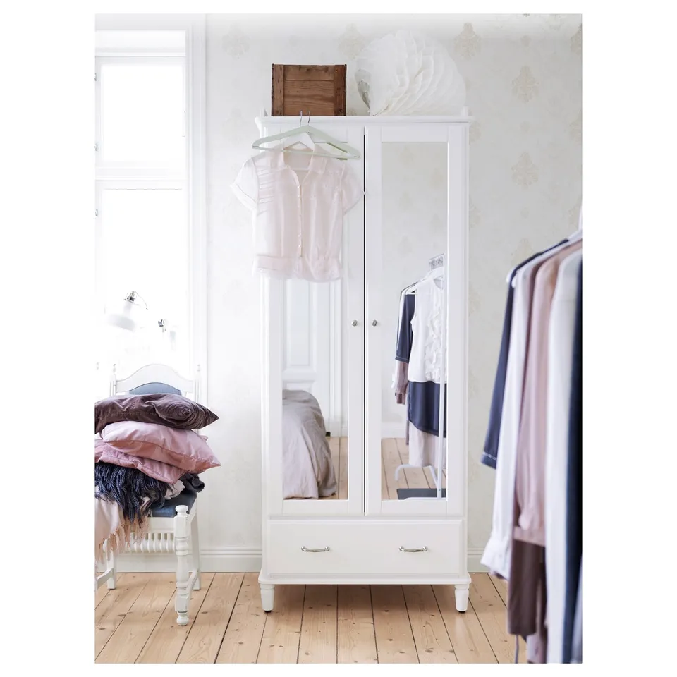 A white TYSSEDAL wardrobe stands in a bright room, mirrored doors reflecting soft pastel clothes and a wood floor.